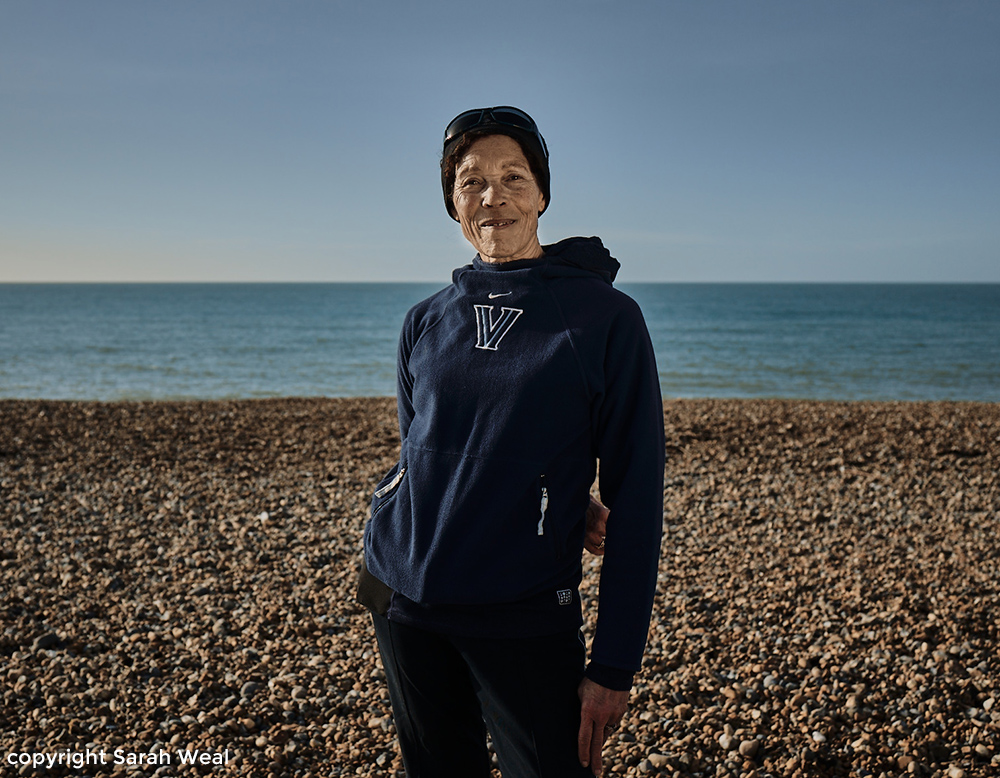 Glenda Jones a beach runner stands on seaford beach with pebbles, and teh blue sea behind her